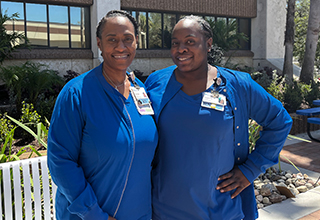 Two Black women dressed in blue scrubs stand arm in arm outside a building.